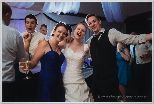 Cayla dances arm in arm between a grinning groomsman and a bridesmaid in blue, guests swaying behind