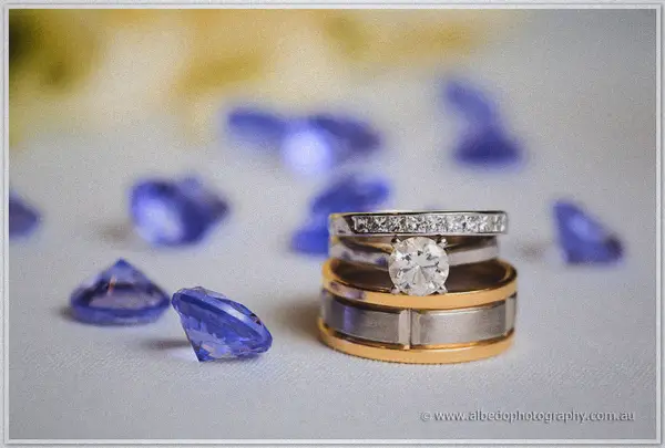 Gold and platinum wedding bands sit with a diamond engagement ring beside scattered sapphire glass gems