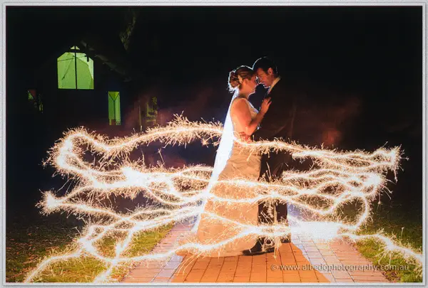 Cayla and Joe embrace at night behind looping sparkler trails on a brick path, resort windows glowing