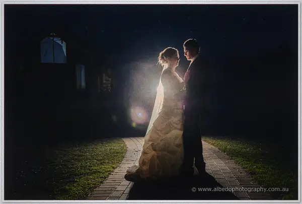 Cayla and Joe stand close on a curved brick path at night, her veil catching the backlight against deep blue