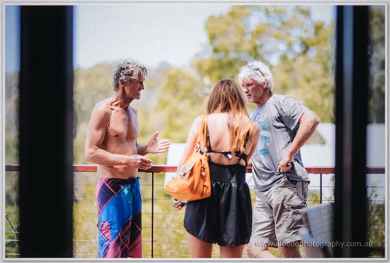 Three guests in board shorts and summer dresses chat on a sun-splashed deck framed by dark posts