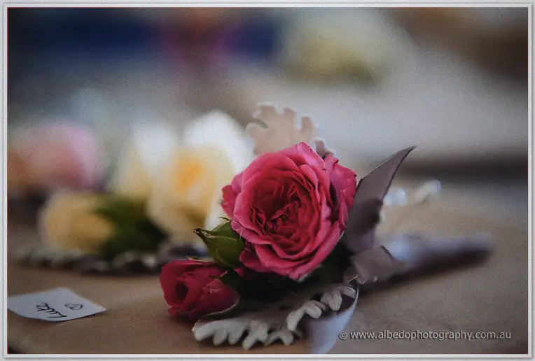 A crimson rose buttonhole with silvery dusty-miller leaf waits on a table beside labelled corsages