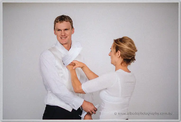 A grinning groom in white vest and tie has his collar adjusted by a smiling helper in white
