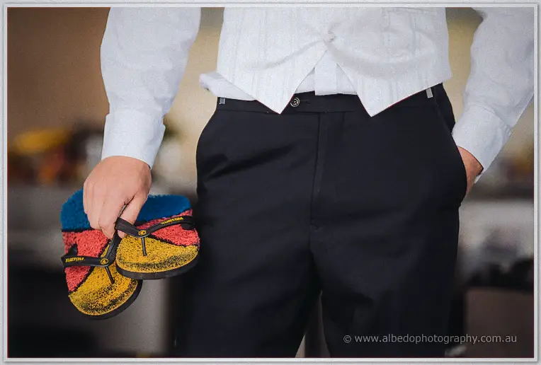 Groom in white vest and black trousers clutches a pair of brightly coloured thongs, hand in pocket