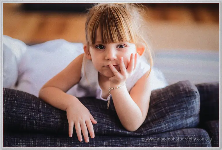 A flower girl in white sprawls on the lounge, chin in hand, gazing at the camera mid-prep