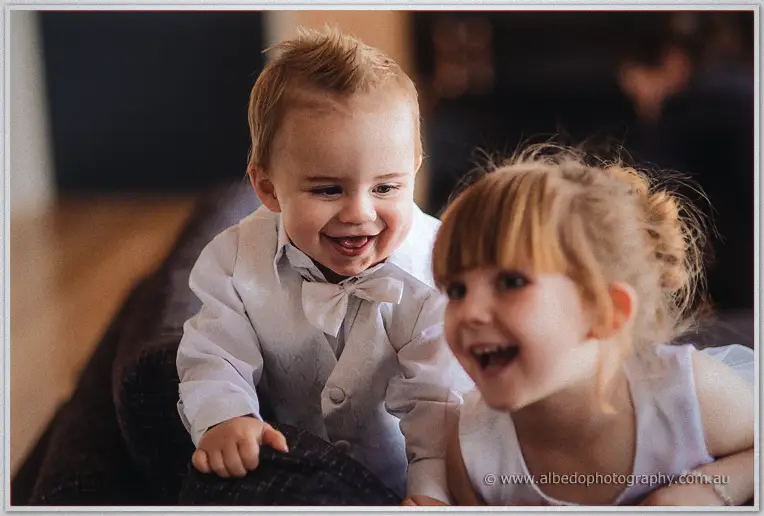 The page boy in a white bow-tied vest and a laughing flower girl tumble together on the couch