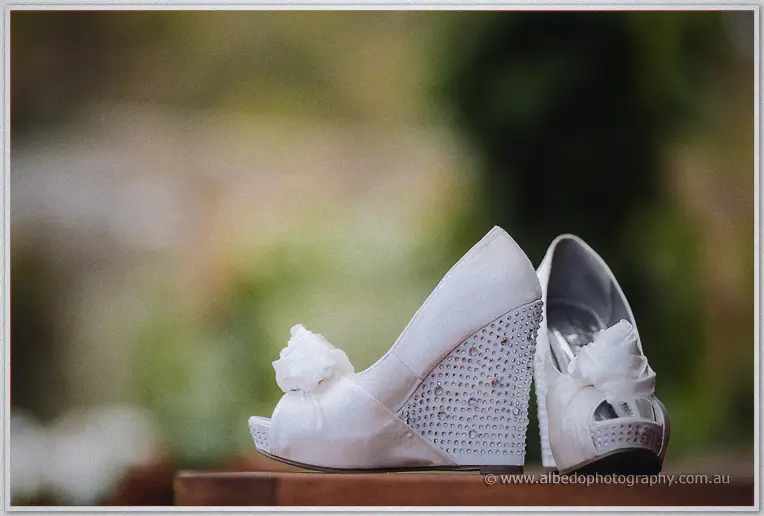 Sparkling white wedge heels with rosette toes sit on a timber rail, greenery washed to a blur