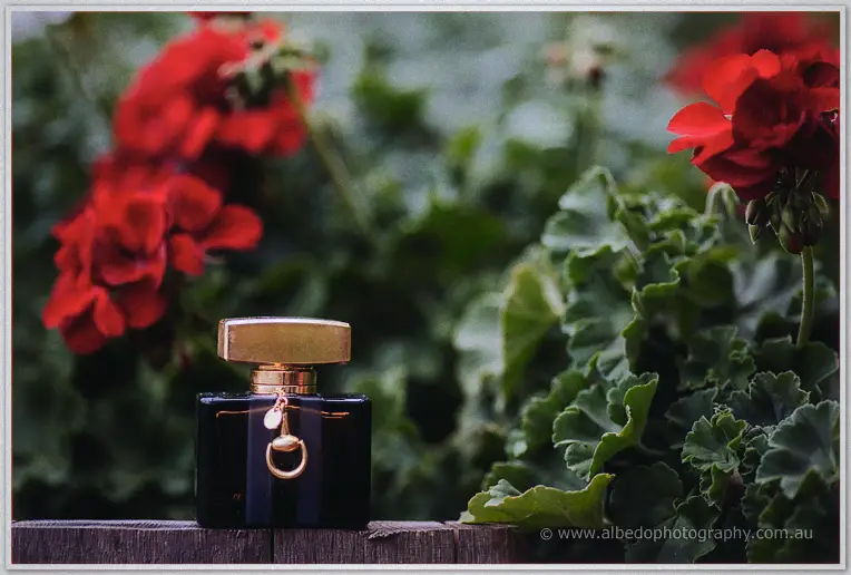A black perfume bottle with gold cap rests on a jarrah railing beside scarlet geraniums