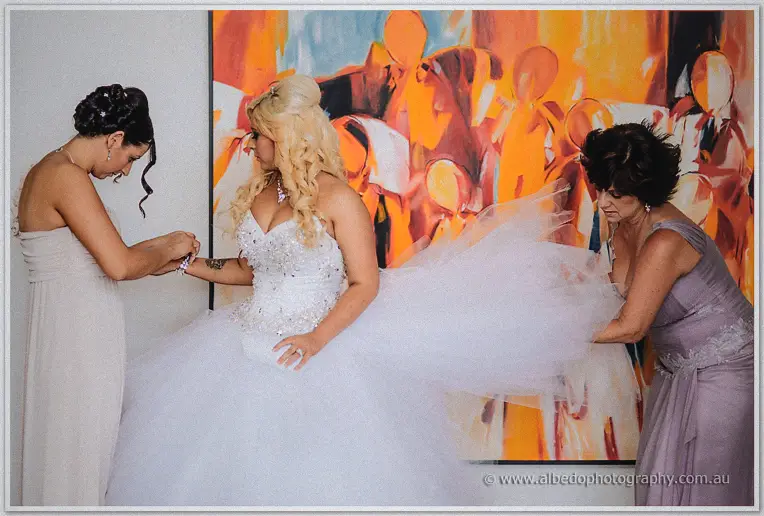 Bridesmaid and mother-in-law-to-be fuss over the bride's veil and tulle train beneath a bold orange artwork