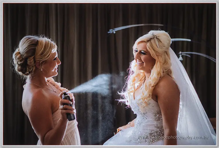 A bridesmaid in blush drenches the veiled bride's curls with hairspray, both laughing in lamp-lit prep