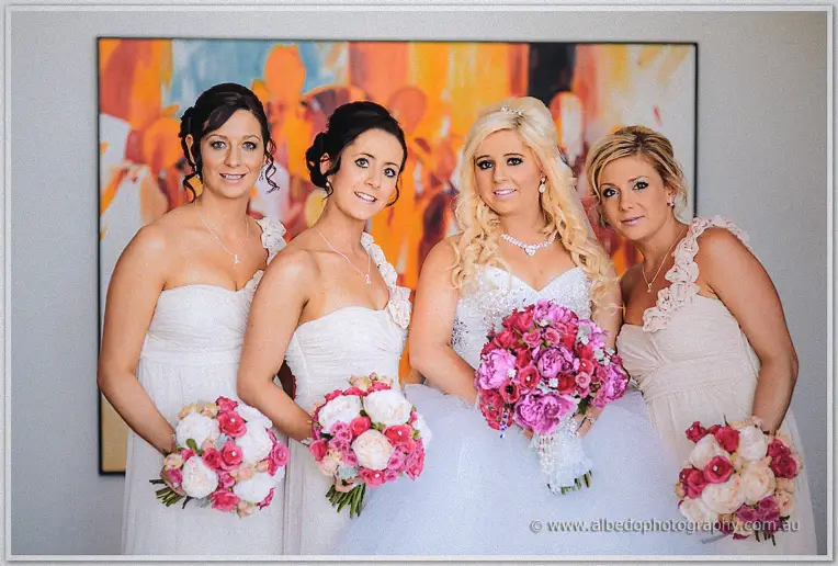 Steph and her bridesmaids in white dresses hold pink bouquets before a vivid abstract painting.