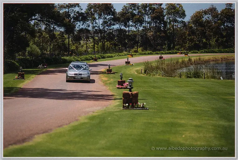 Wedding car with a ribbon bow glides along the tree-lined drive past the lake at Laurance Wines.