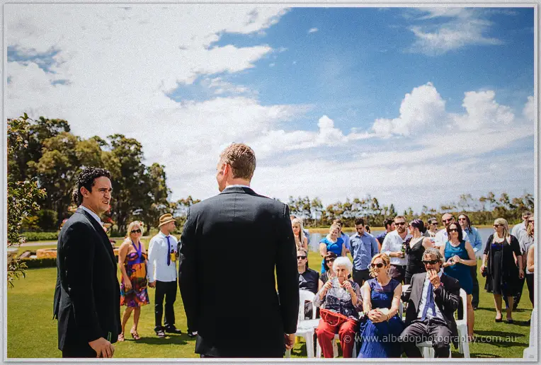 Groom Luke waits with his groomsman as family guests gather on the lawn for the ceremony.
