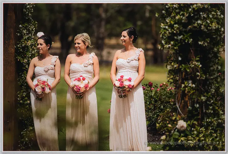 Three bridesmaids in blush gowns holding rose bouquets wait beneath the rose-covered arches.
