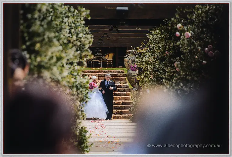 Steph and her father descend the stone steps onto a petal-strewn path through the garden.