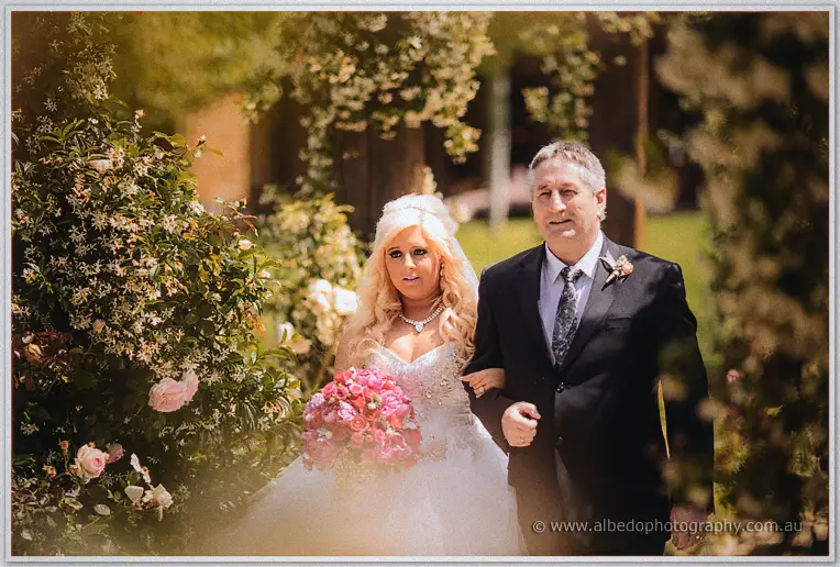 Steph walks arm in arm with her father past pale roses on the sunlit garden aisle.
