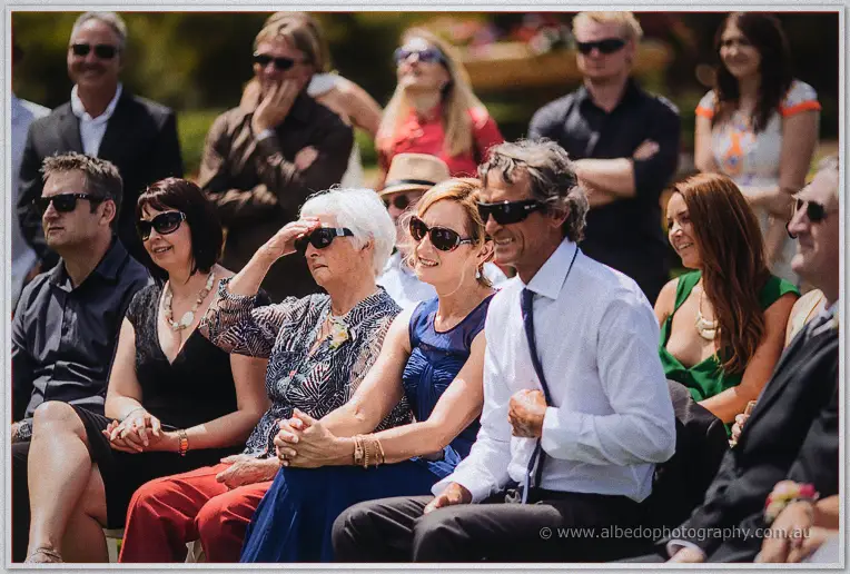 Rows of wedding guests in sunglasses watch the ceremony from white chairs on the lawn.