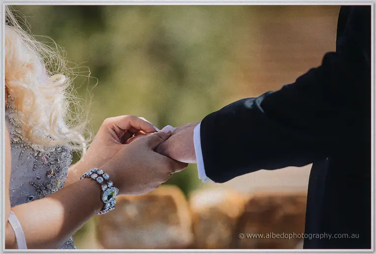 Close-up of Steph's hands placing the wedding ring on Luke's finger in soft garden light.