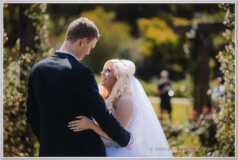 Steph gazes up at Luke as he wraps his arms around her in dappled garden light.