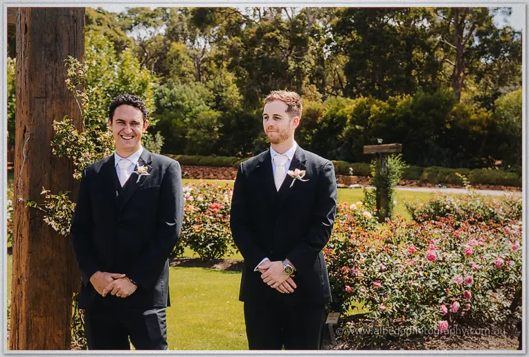 Two groomsmen in dark suits with boutonnieres grin beside the rose beds before the ceremony.