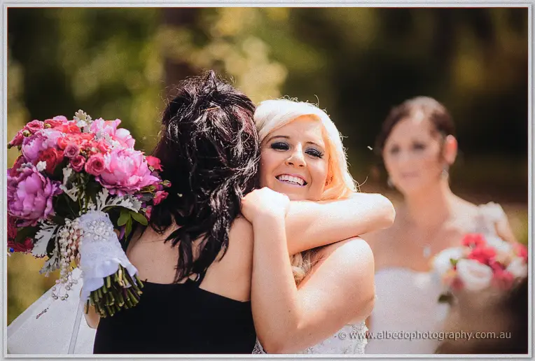 Steph hugs a guest in black on the lawn, bridesmaids with pink bouquets behind her.