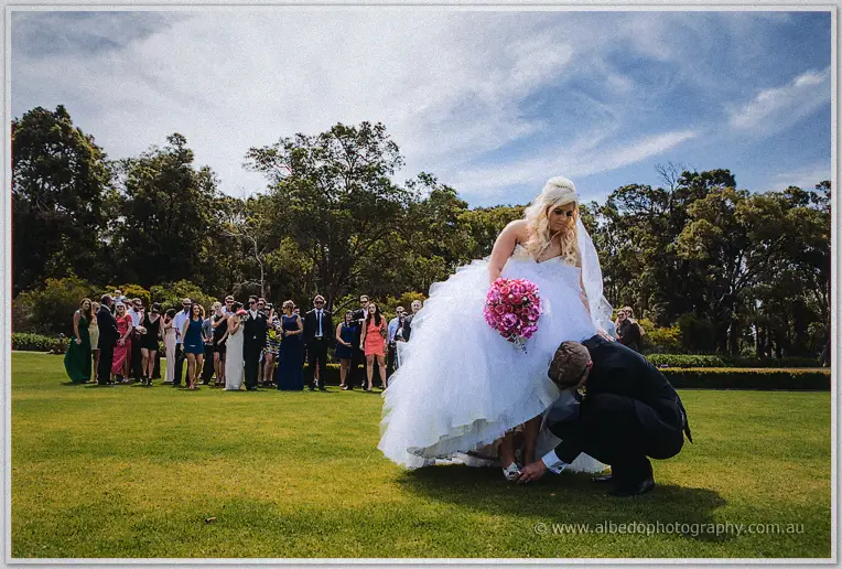Luke kneels to slip Steph's shoe back on as the wedding party watches from the lawn.