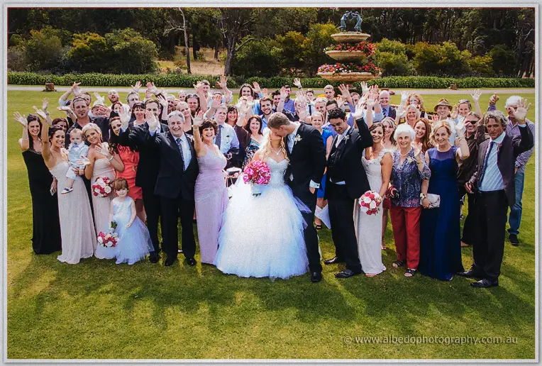 Big group photo of the couple and all their guests waving beneath the garden fountain.