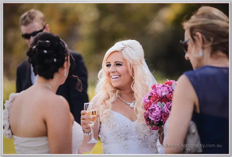 Steph toasts champagne with guests, bouquet in hand, laughing in the afternoon light.