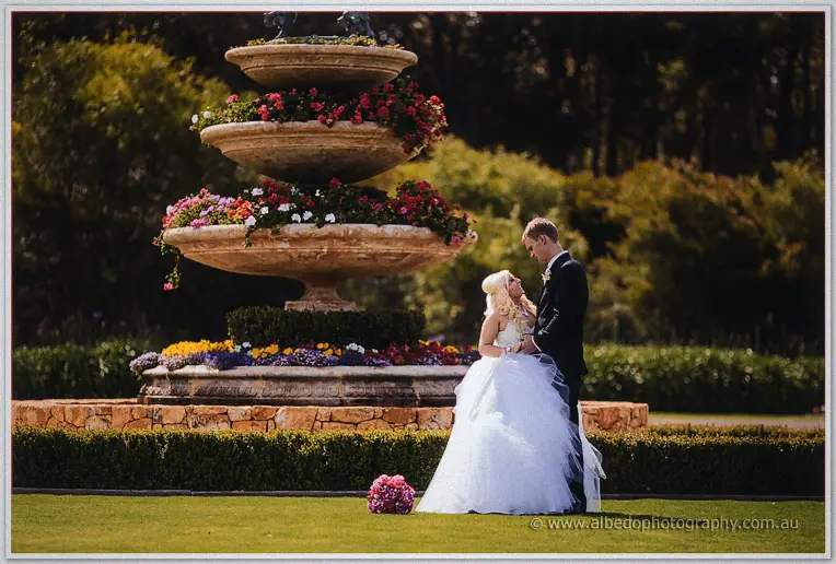 Steph and Luke embrace beside the tiered stone fountain fringed with roses.