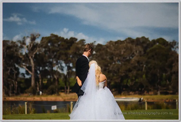 Steph and Luke stand arm in arm looking out over the still lake and gum trees.