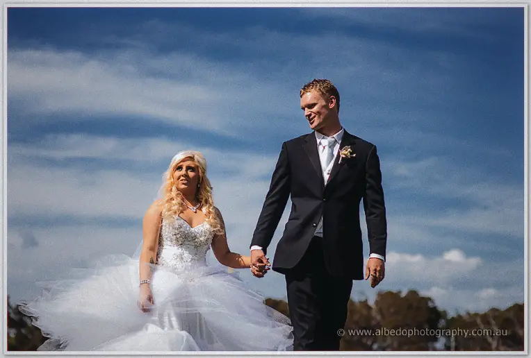 Steph and Luke walk hand in hand across the lawn, tulle dress billowing under a blue sky.