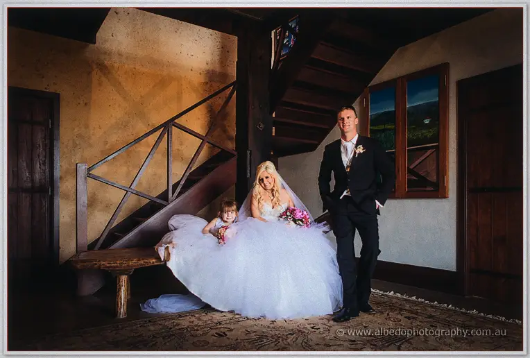 Steph seated with the flower girl by a timber staircase while Luke stands beside her below warm lights.