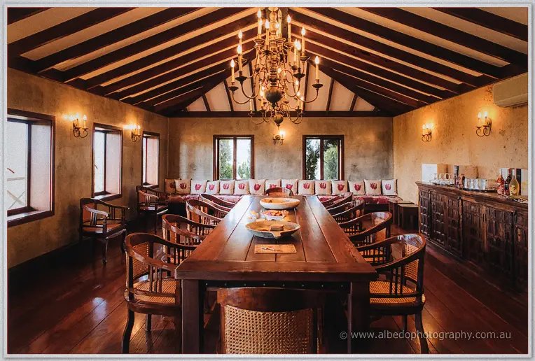 Empty reception room with a long timber table, rattan chairs and candlelit chandelier.