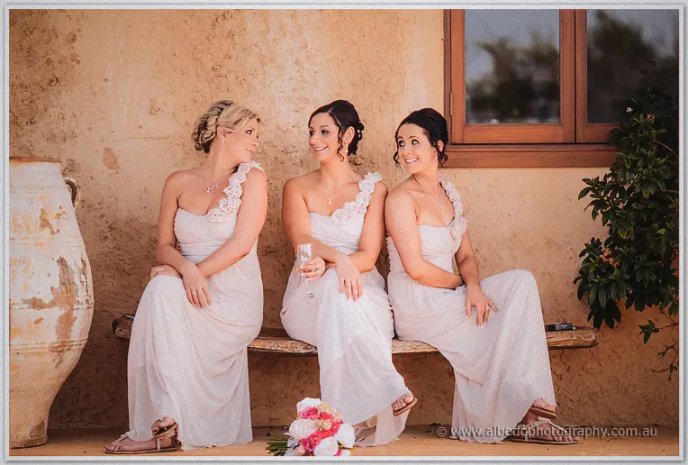 Three bridesmaids in blush gowns sit laughing on a bench against the ochre villa wall.