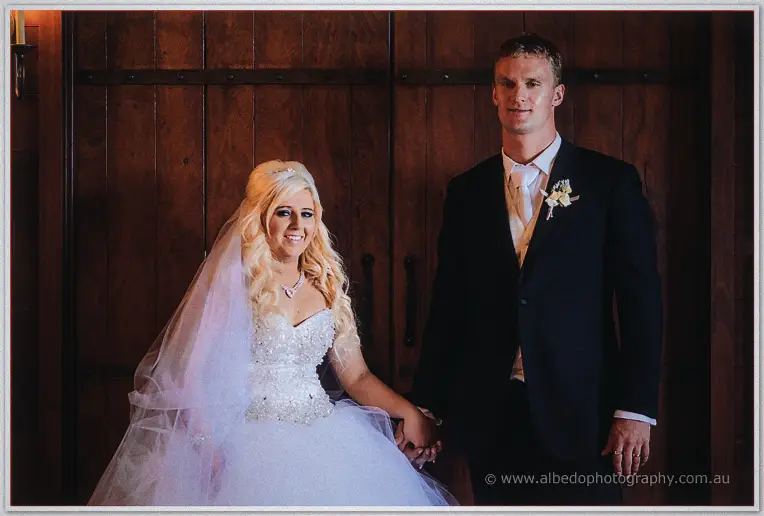 Steph and Luke hold hands before the dark timber doors, her beaded gown catching the light.