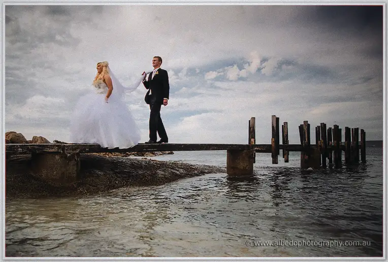 Groom guiding Steph along a broken wooden jetty above calm sea under soft clouds