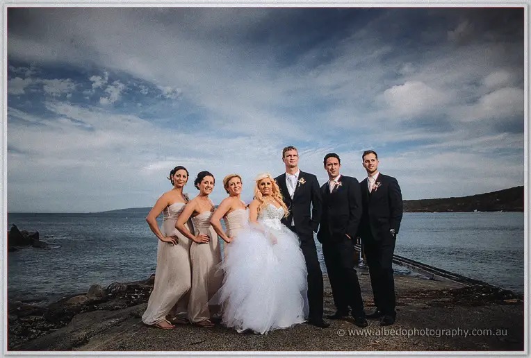 Bridesmaids in blush gowns and groomsmen in black suits grouped with the couple on the rocks