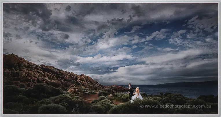 Wide coastal view of the couple dwarfed by granite boulders and moody clouds near Margaret River