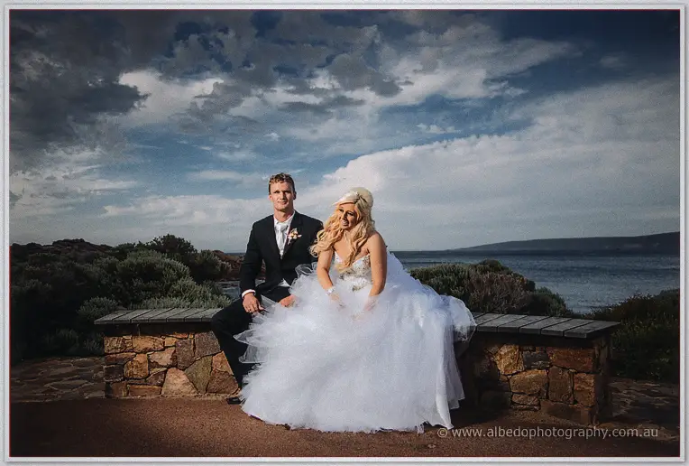Steph and Luke seated on a stone bench above the bay, her gown spread wide around them