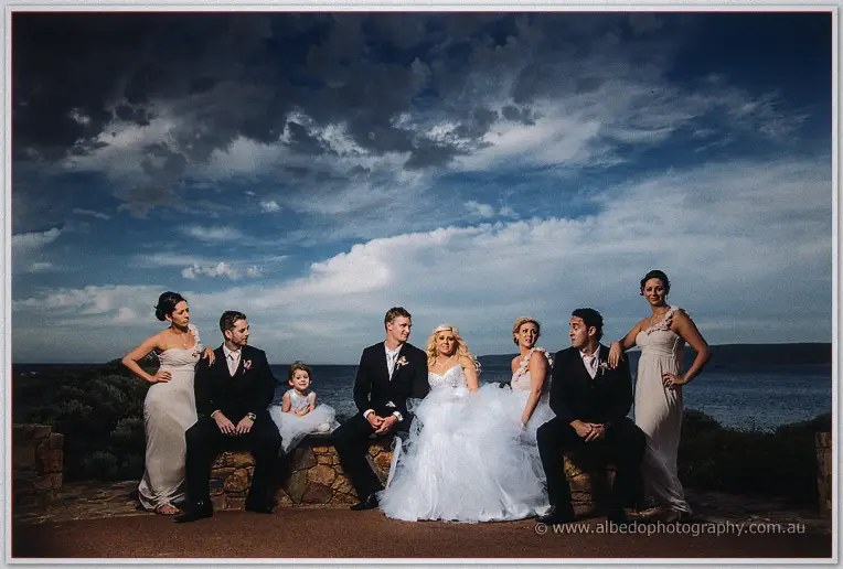 Wedding party seated on a stone wall by the sea, flower girl between the groomsmen
