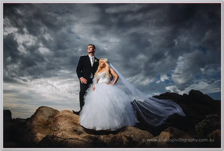 Bride and groom stand on coastal granite rocks beneath a brooding sky, her long veil catching the wind