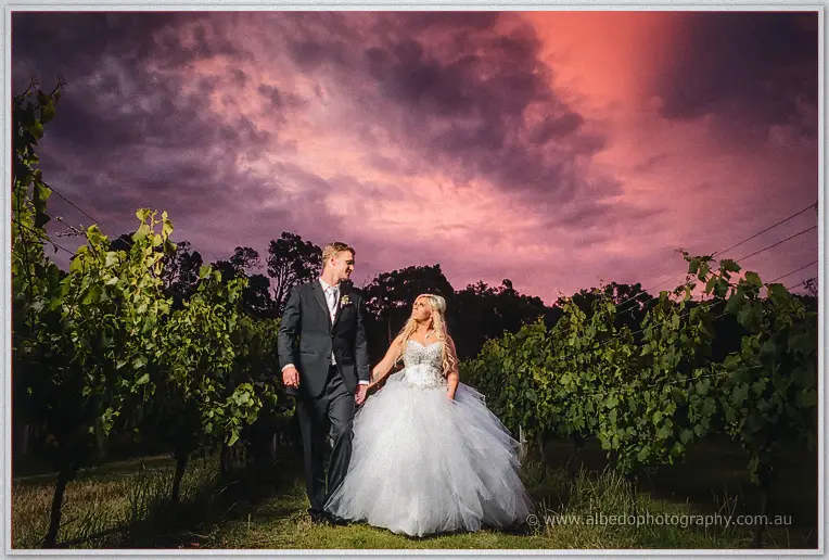 Luke and Steph strolling between vine rows beneath swirling pink and mauve clouds