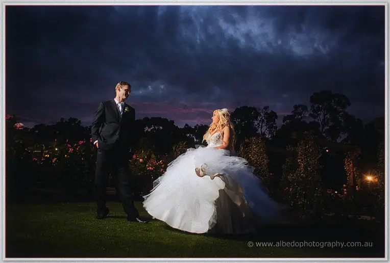 Steph twirling her tulle gown on the lawn as Luke watches beside the rose garden at night