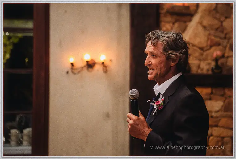 A groomsman in a dark suit delivering a toast into a microphone by candlelight