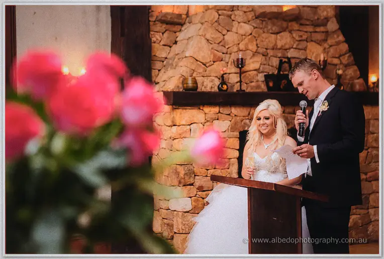 Steph and Luke giving their thank-you speech beside the stone fireplace, roses framing the foreground