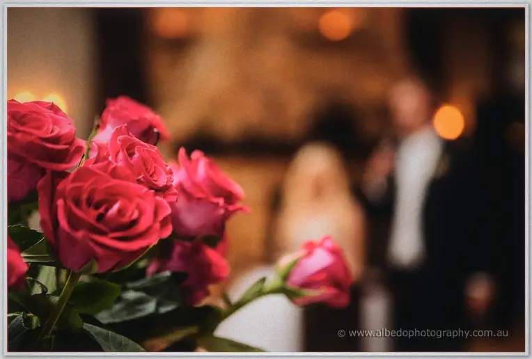 Deep pink roses in sharp focus, the couple blurred mid-speech in the warm background