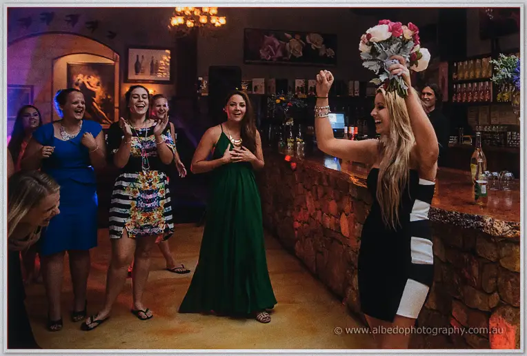 Guest triumphantly raising the caught bouquet as friends cheer around the stone bar
