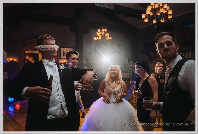 Groom blindfolded with the bride's lace veil as she laughs during a reception game under chandeliers