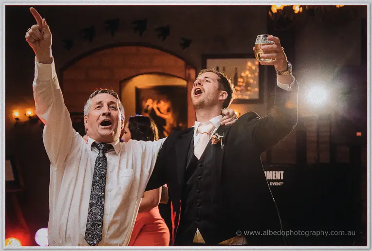 Groom and an older guest sing along with arms raised, drinks in hand, under warm chandelier light