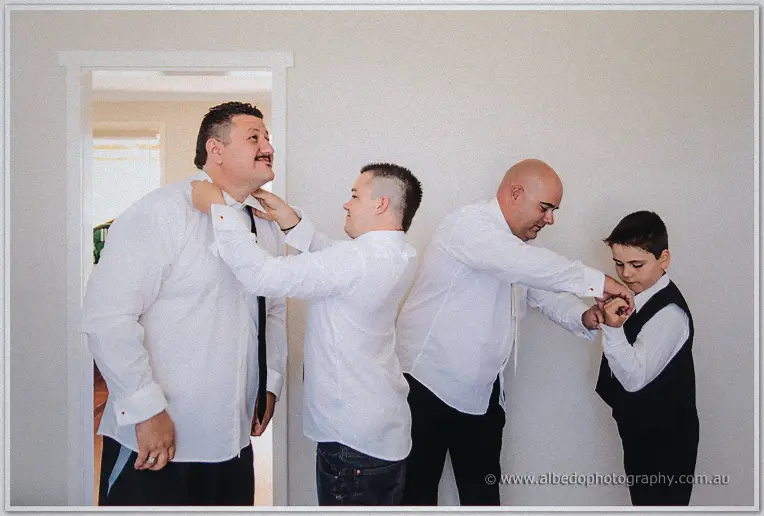 Two men in white shirts knot ties as another helps a small page boy with his cufflink